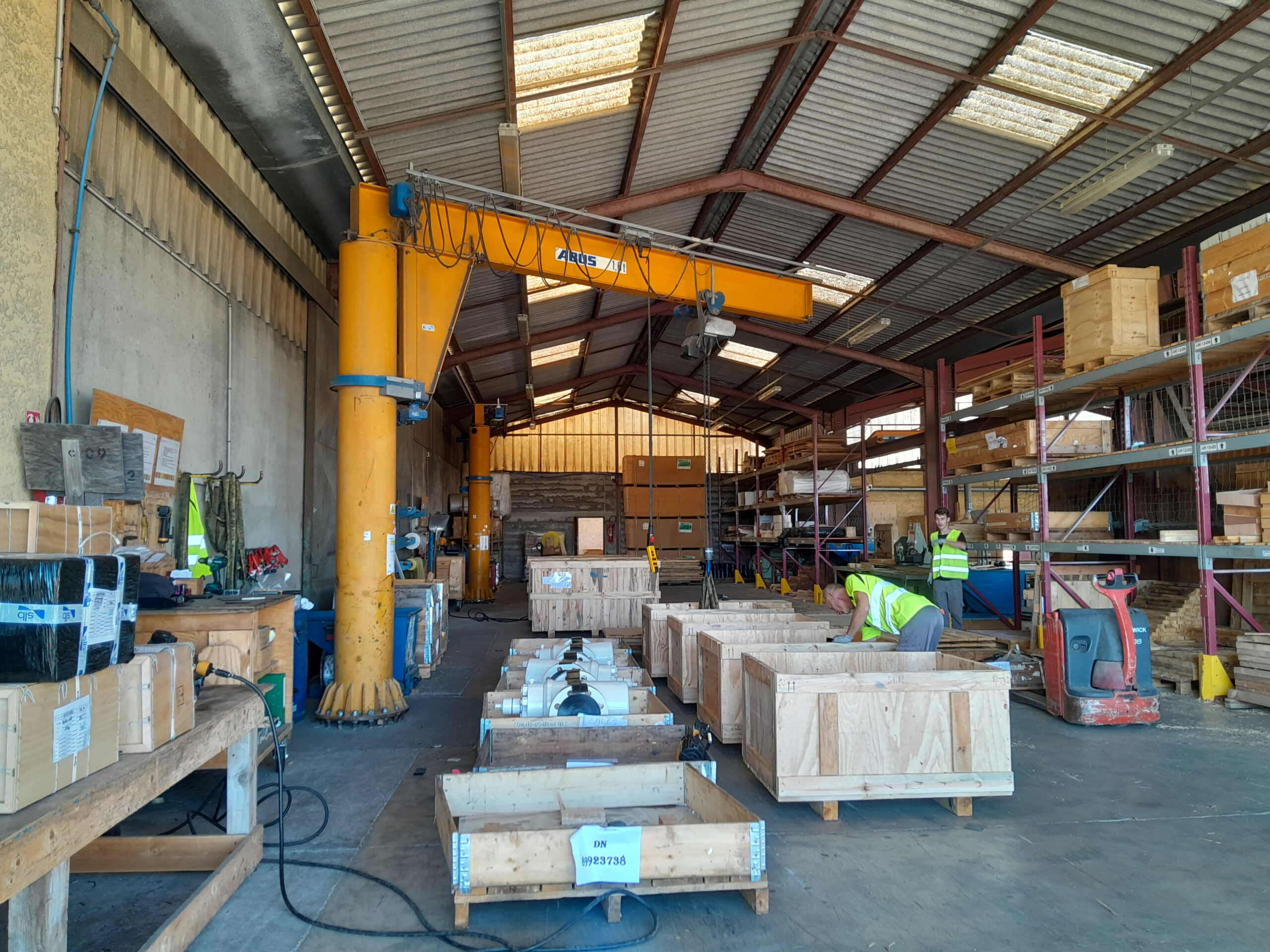 Intérieur d'un atelier industriel avec un pont roulant jaune, des caisses en bois contenant des pièces, et des opérateurs en gilets de sécurité.